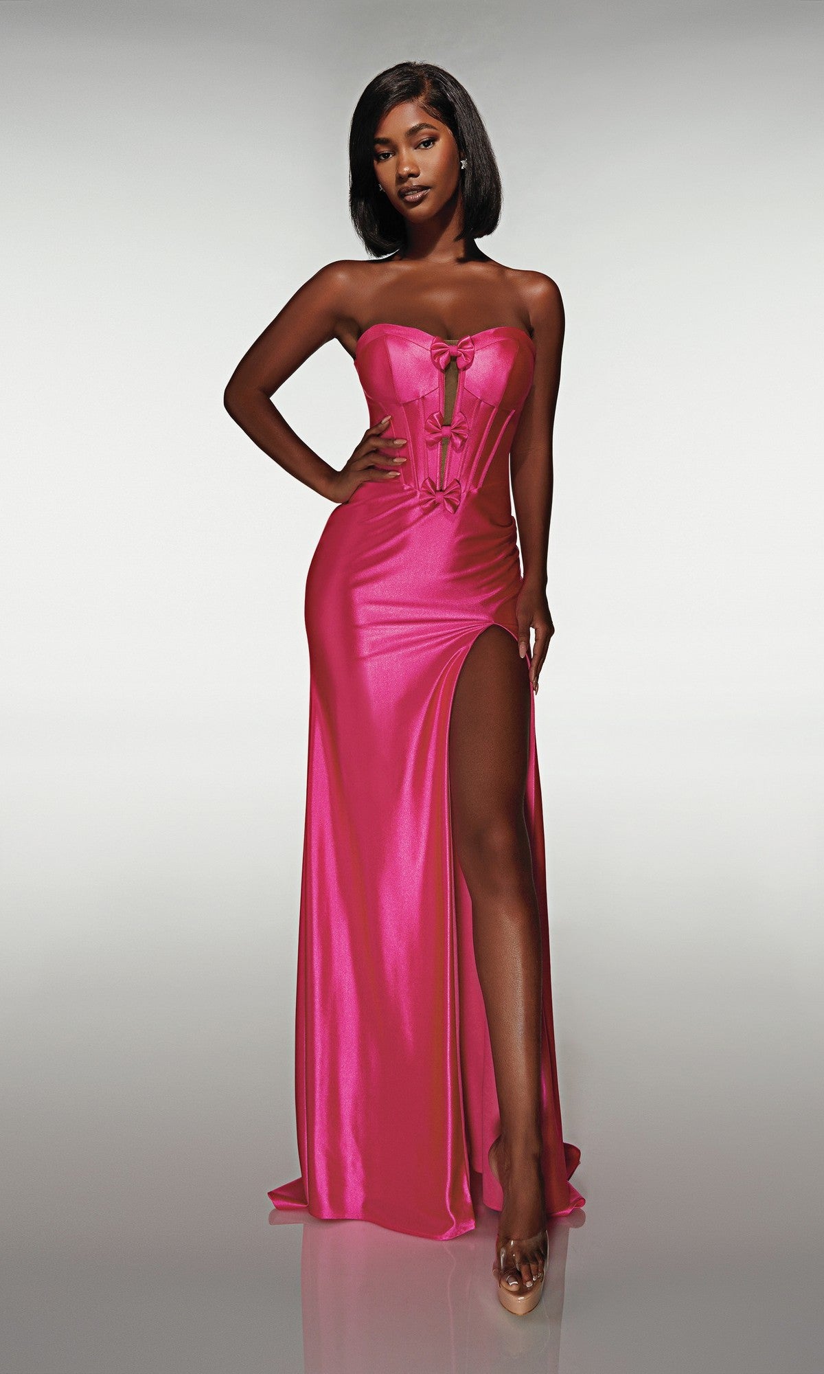 A woman poses against a plain background wearing the Alyce 62046 long prom dress, featuring a strapless corset bodice, floor-length hot pink satin fabric, and a high-leg slit.